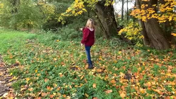 穿着蓝色牛仔裤和一件勃艮第夹克的中年女人在秋天的公园里在黄色和橙色的树间嬉戏视频的预览图