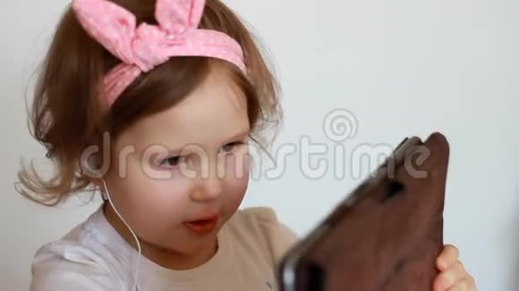一个戴着耳机的小可爱女孩看着屏幕监视器平板电脑看电视录像卡通或玩游戏的滑稽儿童视频的预览图