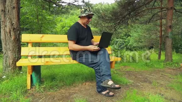 一个戴墨镜的人在公园里坐在长凳上用笔记本电脑工作视频的预览图