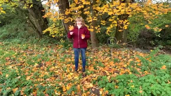 穿着蓝色牛仔裤和一件勃艮第夹克的中年女人在秋天的公园里在黄色和橙色的树间嬉戏视频的预览图