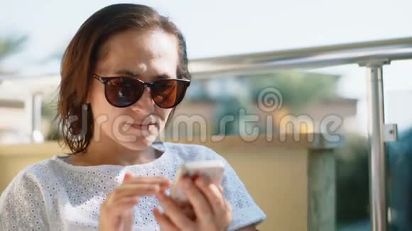 一个戴着智能手机的太阳镜的女人坐在阳台上的玻璃桌旁女人在电话里读短信视频的预览图