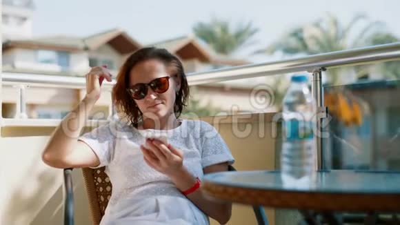 一个戴着智能手机的太阳镜的女人坐在阳台上的玻璃桌旁女人在电话里读短信视频的预览图