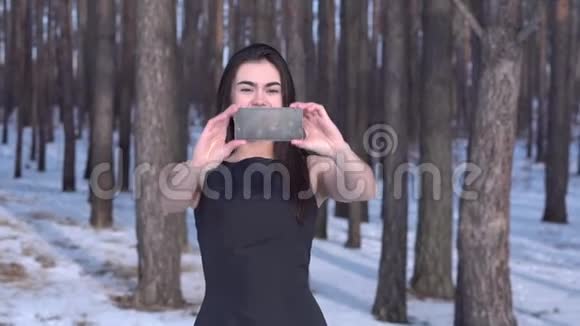 年轻的黑发女孩穿着黑色连衣裙在冬天的森林里微笑着转过身来用智能手机自拍视频的预览图