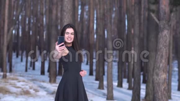 一个年轻的黑发女孩穿着黑色连衣裙一幅迷人的肖像在白雪皑皑的森林景观背景上自拍视频的预览图