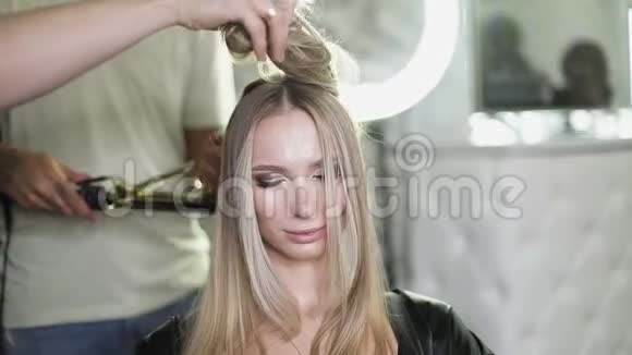 一个男人在美容院用卷发熨斗卷着一个年轻金发模特的头发视频的预览图