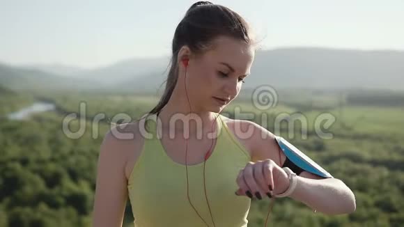 一个穿着运动服的年轻运动女孩在日落时分在山上的新鲜空气中奔跑伴着音乐听着视频的预览图