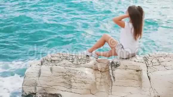 美丽的年轻女孩坐在岩石上挺直了头发享受着清爽的微风和美丽的景色视频的预览图