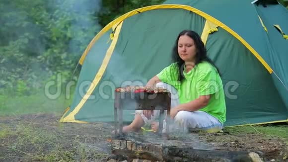 一个年轻的女人坐在一个帐篷背景的森林里在烤架上准备烧烤总体计划视频的预览图