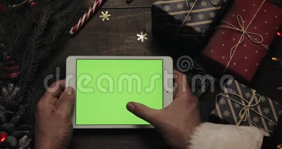 圣诞老人的手在带有绿色屏幕的白色平板电脑上敲击页面另一只手放着一些礼物视频的预览图