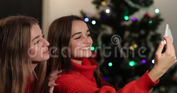 新年庆典4k两个女孩在圣诞树前坐在沙发上的智能手机上自拍视频的预览图
