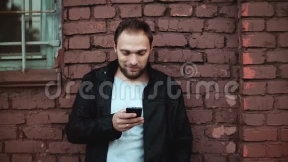 放松的欧洲男人在外面使用智能手机应用程序英俊的胡子男商人移动办公室红砖墙4K视频的预览图