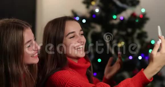 新年庆典4k两个女孩在圣诞树前坐在沙发上的智能手机上自拍视频的预览图