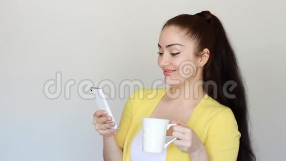 一个美丽的黑发女孩的肖像她从杯子里喝了一杯热饮料手里拿着一部手机青年视频的预览图