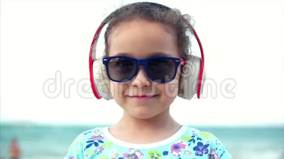 海滩上快乐的小女孩戴着太阳镜听着耳机上的音乐看着相机微笑着还有一个视频的预览图