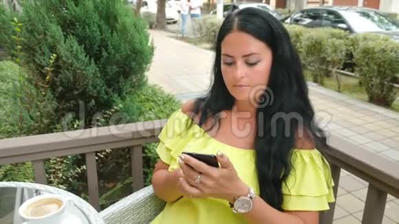 年轻漂亮的黑发女人坐在户外咖啡馆里喝咖啡用电话视频的预览图
