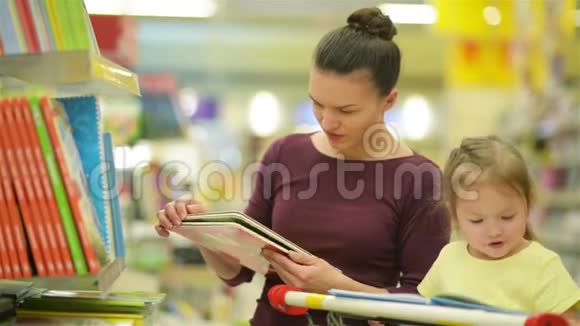 年轻的母亲和她的女儿在超市选书美丽的女儿坐在超市的购物车里视频的预览图