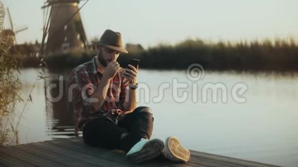 4K成功的欧洲农民坐在湖边码头上男人穿着格子衬衫和帽子使用智能手机移动购物应用程序视频的预览图