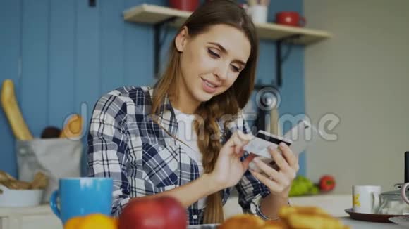 在家里的厨房里吃早餐时一位快乐的女士用智能手机和信用卡在线支付账单视频的预览图