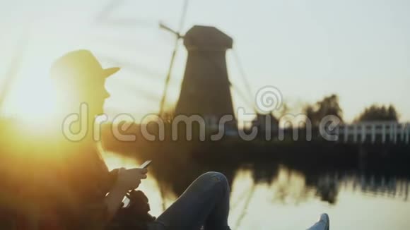 千禧女孩坐在湖边码头打电话夕阳西下女人在户外使用购物应用程序古老的荷兰风车4K视频的预览图