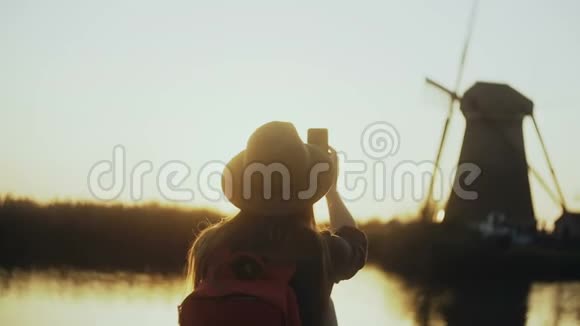 戴帽子的女人坐在夕阳下的乡村湖码头上女孩用她的智能手机拍了一张旧风车照片4K后视图视频的预览图