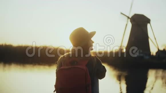 戴帽子的女人坐在宁静的日落湖码头上看上去不错拿着她的手机传统的荷兰风车4K后视图视频的预览图