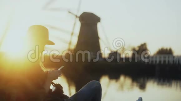 戴帽子的女孩坐在夕阳下的乡村湖码头上旅游妇女用智能手机拍一张荷兰旧风车的照片4K视频的预览图