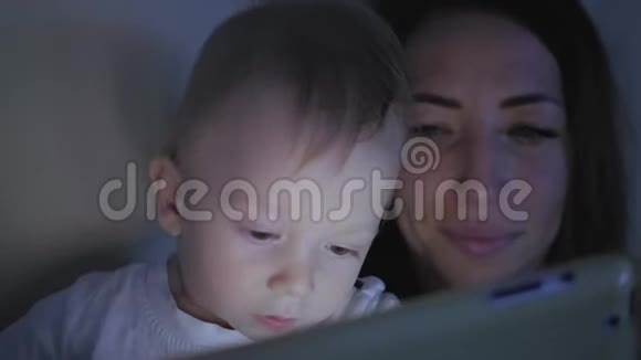晚上妈妈和小儿子坐在家里的沙发上玩数字平板电脑视频的预览图