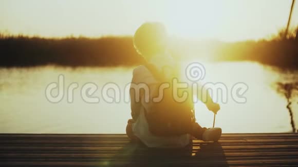 男孩在湖边码头玩棍子后景带背包的小孩坐在码头上惊人的太阳镜头耀斑4K视频的预览图