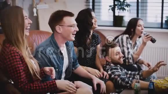 朋友们坐在沙发上在电视上看有趣的电影白人年轻人坐在沙发上喝饮料和零食电影之夜视频的预览图