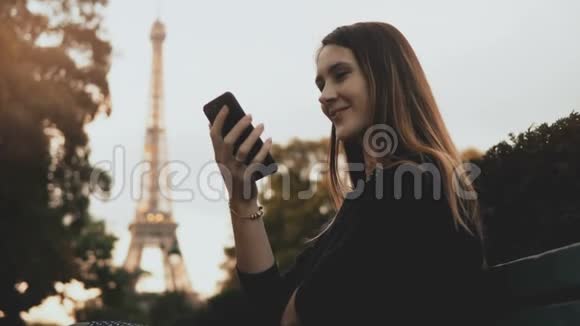 年轻快乐的女人独自坐在法国巴黎埃菲尔铁塔附近的长凳上使用智能手机视频的预览图