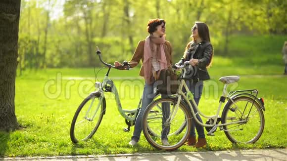 两个女朋友站在公园里拿着自行车互相交谈背景上的绿草视频的预览图