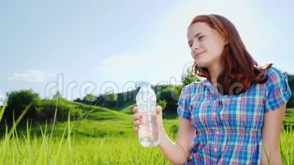 一个13岁的女孩从塑料瓶里喝水在风景如画的绿色草地上野餐视频的预览图