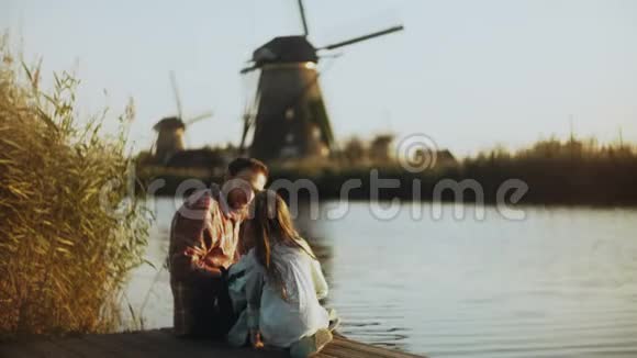 单身父亲和两个孩子坐在阳光明媚的湖码头上荷兰传统风车家庭关系4K视频的预览图