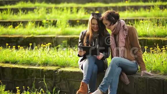 两个穿牛仔裤的可爱女孩正在公园里使用平板户外活动年轻迷人的女人正在看视频的预览图