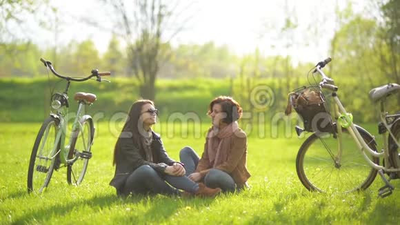 漂亮的女朋友在靠近自行车和树木的公园里在绿草上坐着一个会话门视频的预览图