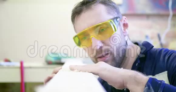 木工车间的一名男子戴着防护眼镜用手从木屑中清理木板并检查视频的预览图