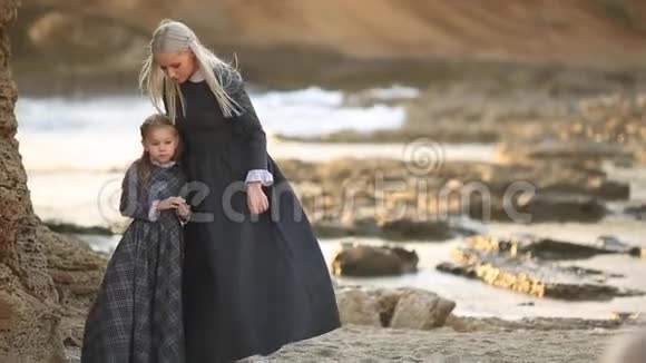 美丽的母亲金发穿着一件灰色的长裙子她的小女儿穿着海滩上的裙子视频的预览图