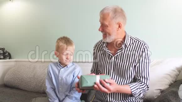 一代人祖父和孙子带着礼品盒坐在家里的沙发上视频的预览图