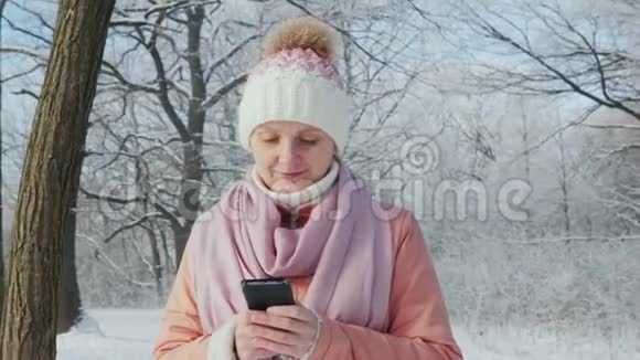 年轻女子在冬季公园散步享受智能手机视频的预览图