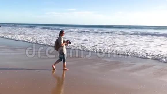 一位女摄影师赤脚走在海滩上停下来拍一张照片视频的预览图