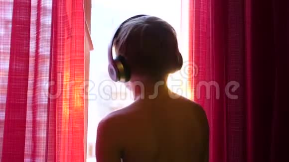 年轻人站在窗边跳舞通过耳机听音乐背景模糊日落视频的预览图