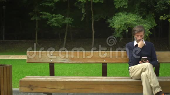 中年男人坐在长凳上使用智能手机聊天在公园里休息视频的预览图