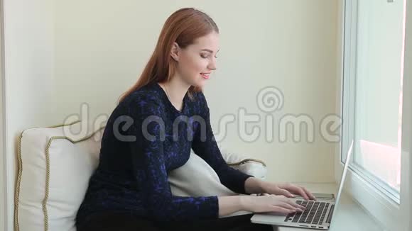 漂亮的年轻女性在家坐在窗台上用笔记本电脑在社交网络上交流视频的预览图