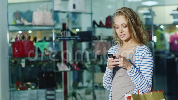 漂亮的女购物者带着购物袋使用智能手机在玻璃橱窗鞋店对面视频的预览图