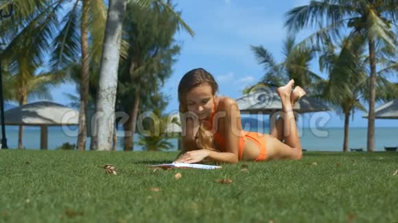 穿泳装的漂亮女孩在棕榈树的草地上看书视频的预览图