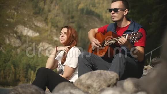 一个戴墨镜的年轻人在阳光明媚的阿尔泰共和国山河边的岩石上弹吉他视频的预览图
