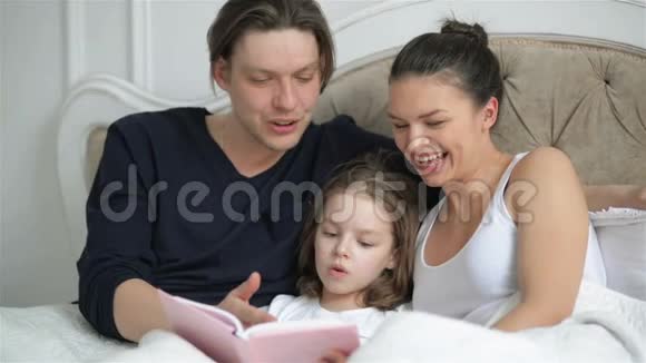 聪明的小女孩正在读一本躺在床上的书她年轻的父母在帮助她女童学习视频的预览图