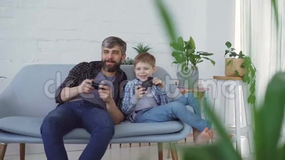 父子俩坐在家里的沙发上玩电子游戏儿子赢了感到兴奋笑了视频的预览图