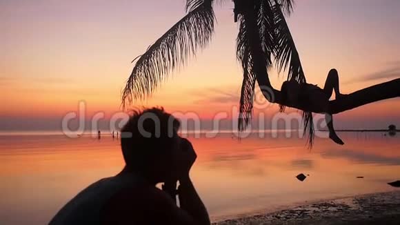 海滩上年轻夫妇的剪影男人拍他女朋友的照片而女孩则躺在棕榈树上视频的预览图