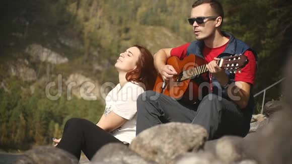 一个戴墨镜的年轻人在阳光明媚的阿尔泰共和国山河边的岩石上弹吉他视频的预览图